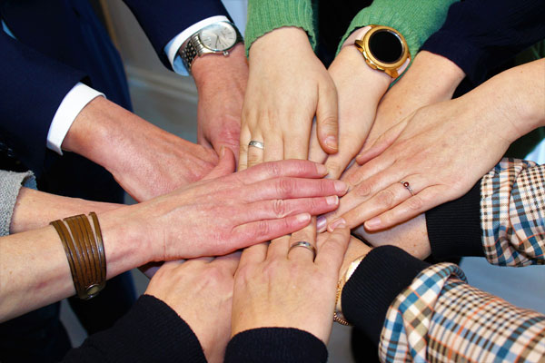 Eine Gruppe von Menschen reicht sich in der Mitte die Hände und zeigt damit Einigkeit und Teamwork. Verschiedene Uhren, Ringe und Kleidungsstile sind zu sehen und unterstreichen die Vielfalt.