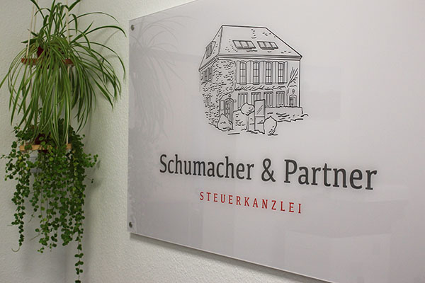 Auf einem Schild an der Wand steht Schumacher & Partner STEUERKANZLEI, darüber ist ein Gebäude abgebildet; daneben steht eine grüne Topfpflanze.